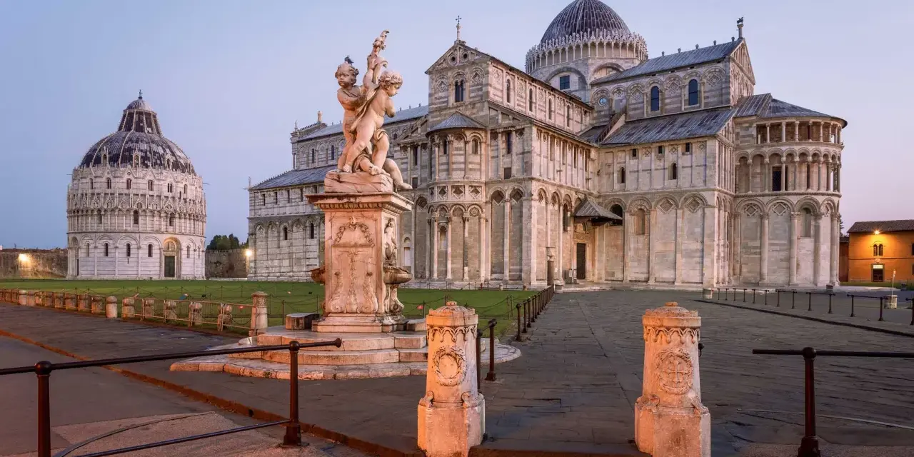 Il **Battistero di Pisa** e il Duomo illuminati dalla luce dell'alba. Una fontana con putti al centro della piazza.