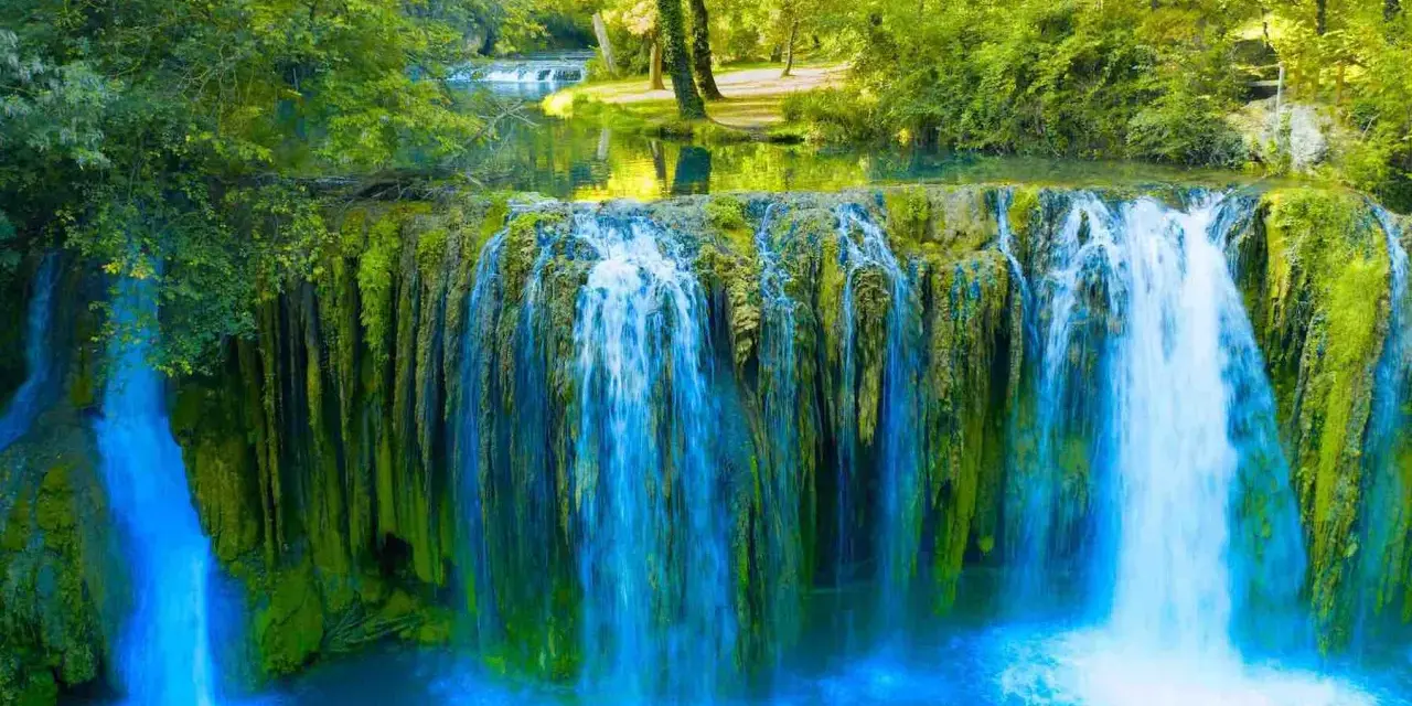 Cascata rigogliosa nel Val d'Arno, con acqua azzurra che scende su rocce ricoperte di muschio verde.