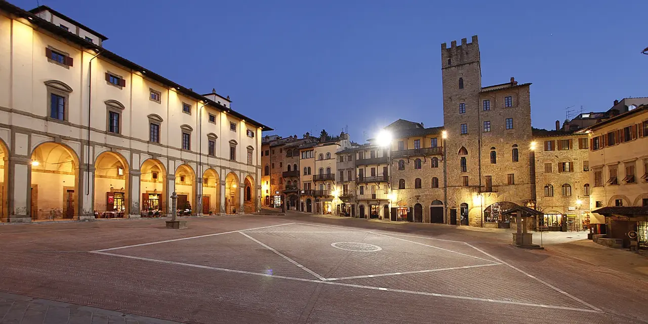 Piazza illuminata di Arezzo al crepuscolo, con portici, negozi e una torre medievale. Qui la vita è bella.
