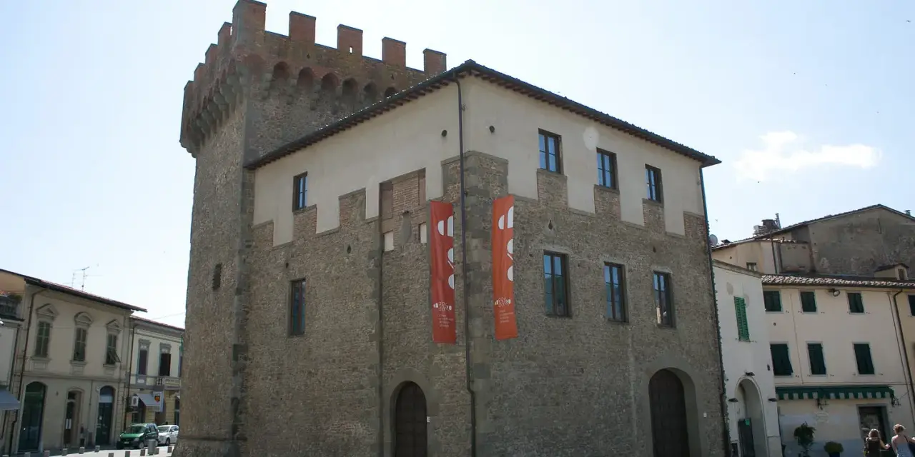 Il cassero di Montevarchi, un'antica torre con merlature, si erge imponente accanto a edifici storici, con bandiere rosse che adornano la facciata.