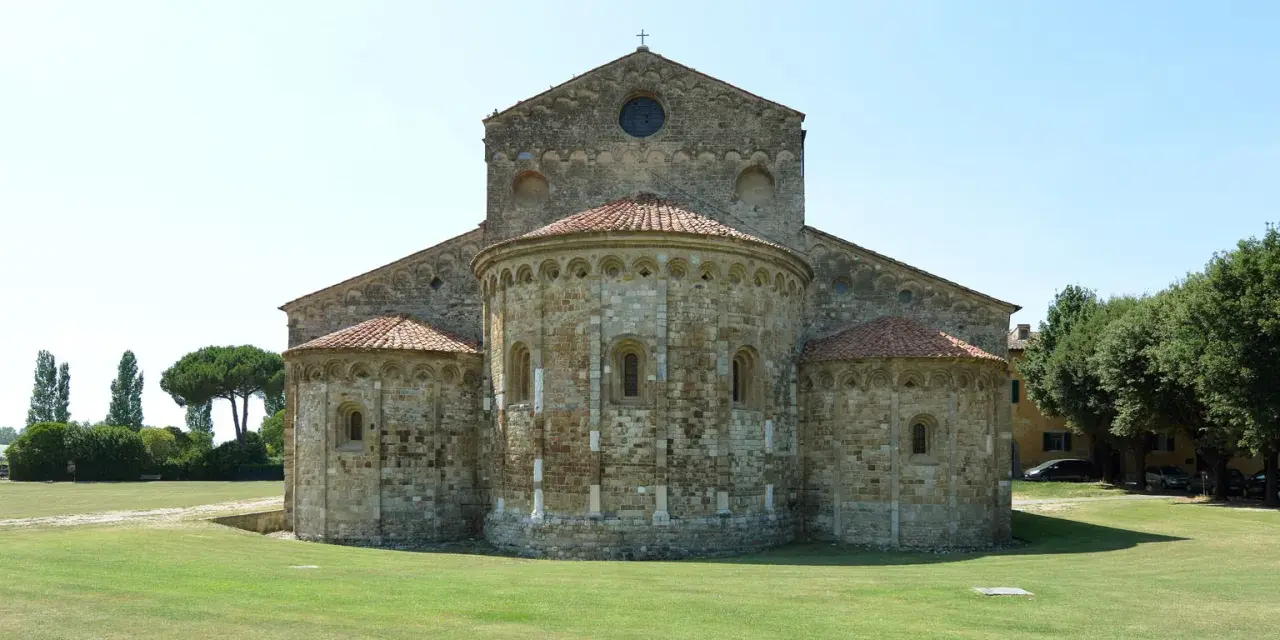 La Basilica di San Piero a Grado, con le sue absidi romaniche e il prato verde circostante, si staglia contro un cielo limpido.