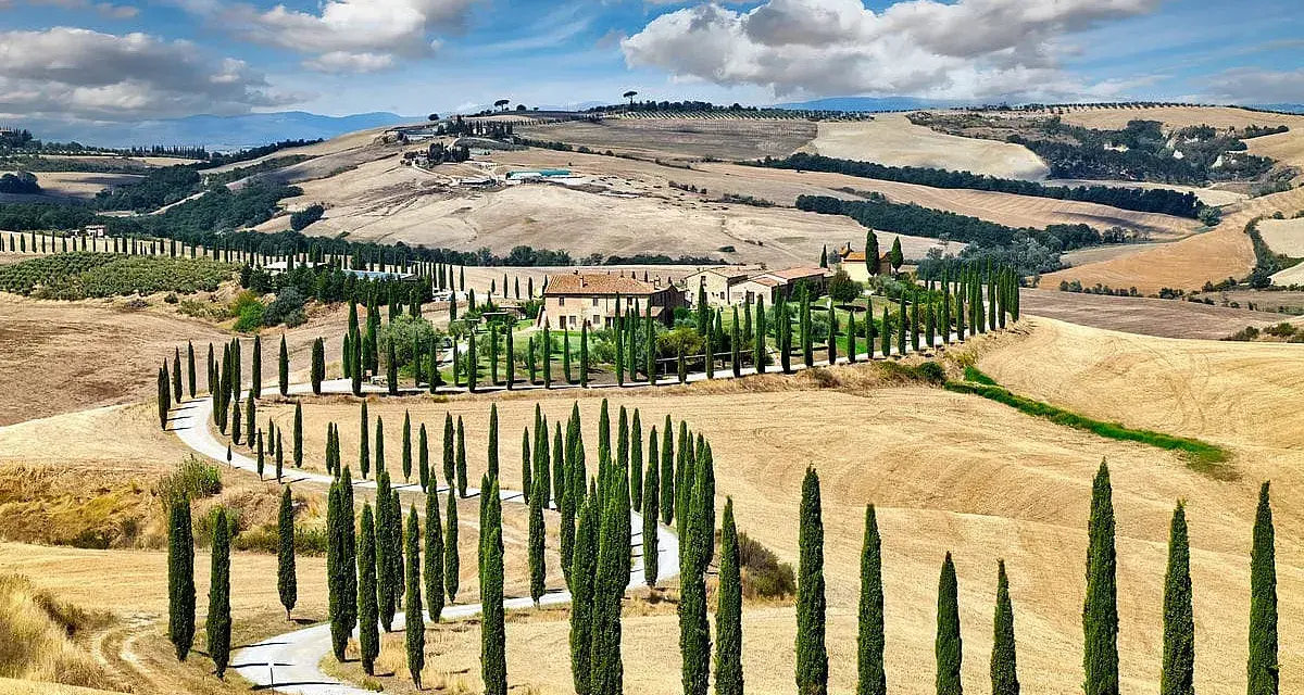 Un casale nel Val d'Orcia, circondato da cipressi e dolci colline dorate sotto un cielo azzurro con nuvole bianche.