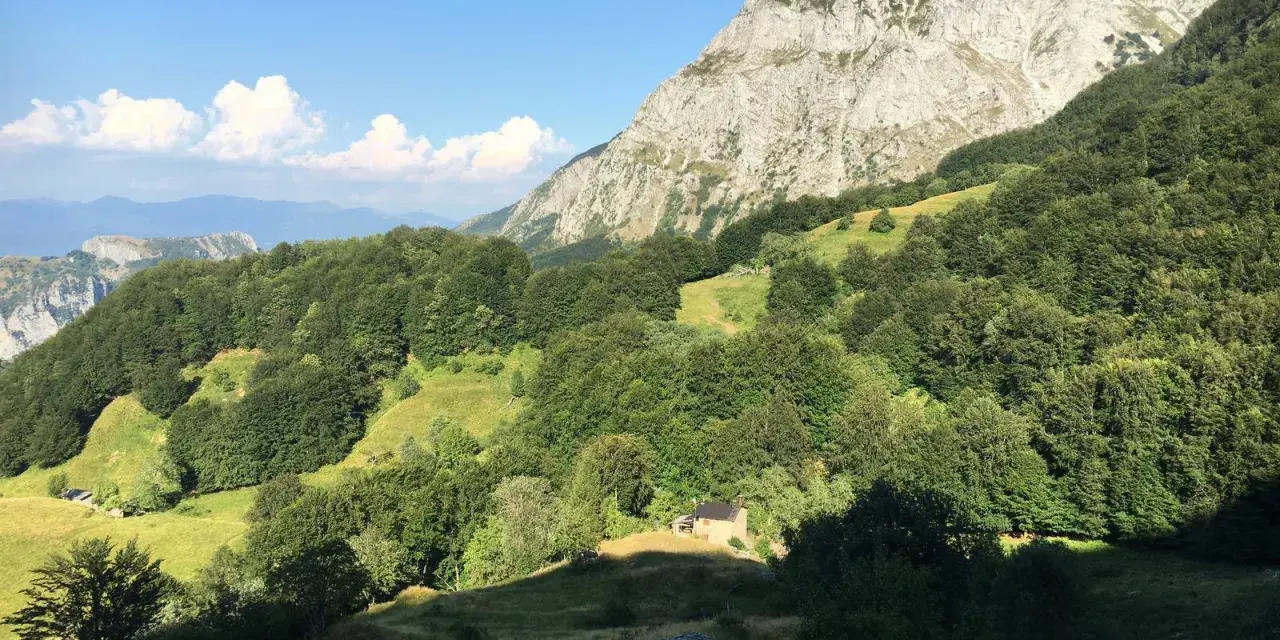 Un piccolo rifugio solitario ai piedi del **Puntato Apuane**, circondato da boschi verdi e montagne imponenti sotto un cielo azzurro.