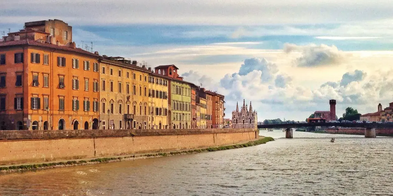 Edifici colorati lungo il fiume Arno a Pisa, con un ponte e nuvole suggestive nel cielo. Un'immagine che cattura la bellezza di Pisa e dintorni.