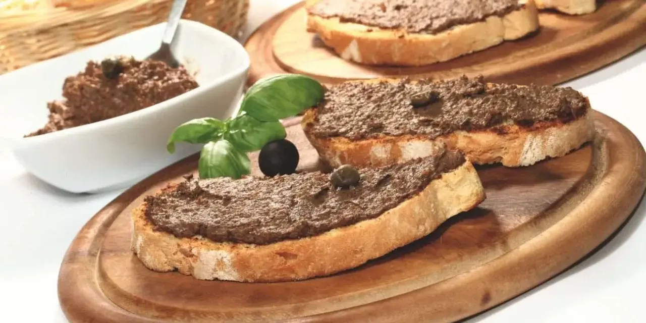 Toasted bread slices topped with dark spread, garnished with basil, olives, and capers.