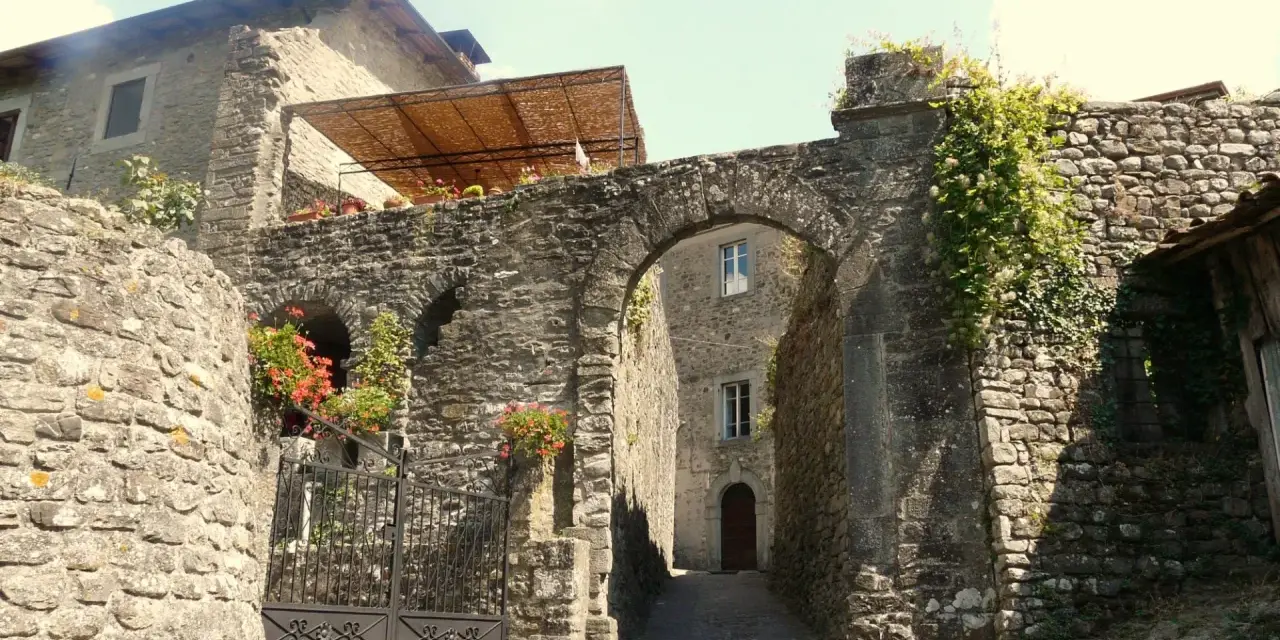 Un vicolo antico a Casola in Lunigiana, con un arco in pietra, mura di sassi e fiori rossi che sbocciano.