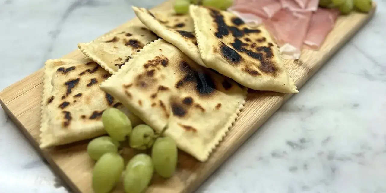 Tortelli alla lastra casentino, dorati e leggermente bruciacchiati, serviti con uva fresca e affettati di prosciutto su un tagliere di legno.