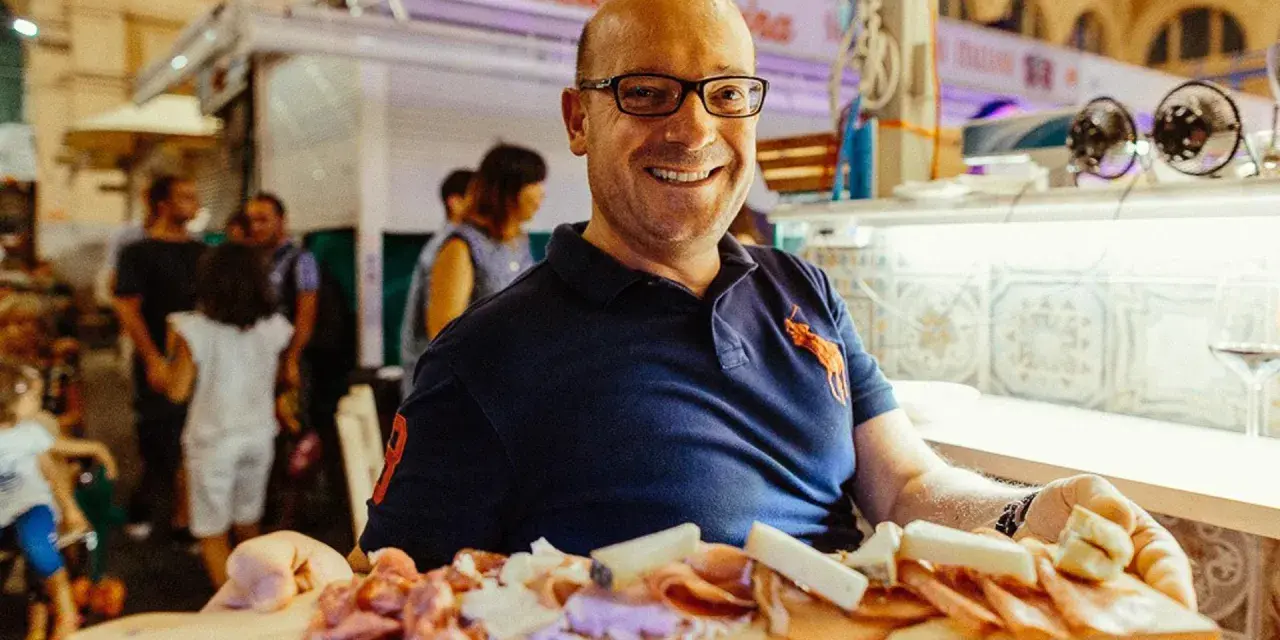 Uomo sorridente al mercato delle vettovaglie Livorno, offre un tagliere di salumi e formaggi.