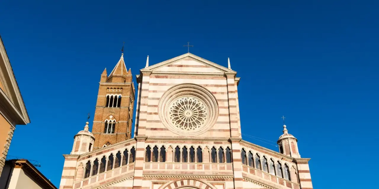 Il Duomo di Grosseto, con la sua facciata a strisce bianche e rosa e il campanile in mattoni, si staglia contro un cielo azzurro intenso.