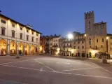 Piazza illuminata di Arezzo al crepuscolo, con portici, negozi e una torre medievale. Qui la vita è bella.
