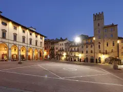 Piazza illuminata di Arezzo al crepuscolo, con portici, negozi e una torre medievale. Qui la vita è bella.