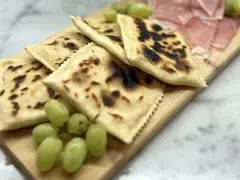 Tortelli alla lastra casentino, dorati e leggermente bruciacchiati, serviti con uva fresca e affettati di prosciutto su un tagliere di legno.