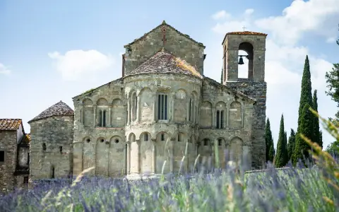 La Pieve di Romena, un'antica chiesa in pietra con campanile, si erge maestosa tra cipressi e un campo fiorito di lavanda sotto un cielo azzurro.
