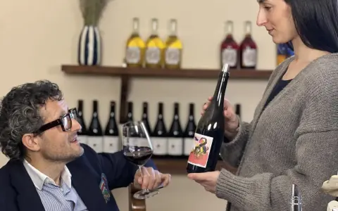 Uomo con occhiali e bicchiere di vino rosso, donna presenta bottiglia. Degustazione vini Arezzo in cantina.