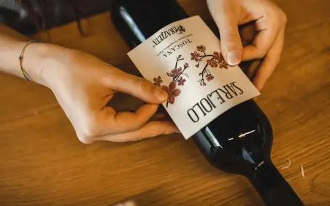 Mani che applicano un'etichetta su una bottiglia di vino. L'etichetta indica "Toscana", la regione di origine del vino Sangiovese.