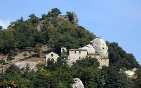 Antico eremo di Cerbaiolo, un complesso di edifici in pietra arroccato su un colle boscoso, con una croce sulla cima di una roccia.