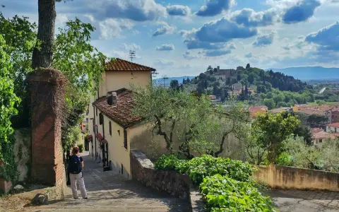 Una persona scende una scalinata in pietra, con case antiche e vegetazione rigogliosa. In lontananza, il **monte Morello** domina il paesaggio collinare.