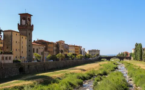 Pescia: cosa vedere? Un fiume serpeggia tra case colorate e un'antica torre con orologio, sotto un cielo azzurro.