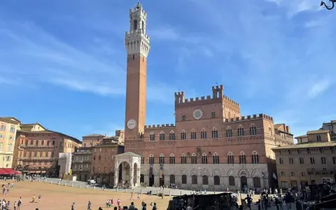 La Piazza del Campo di Siena, con la Torre del Mangia che domina il cielo azzurro, brulica di gente che passeggia e si gode il sole.