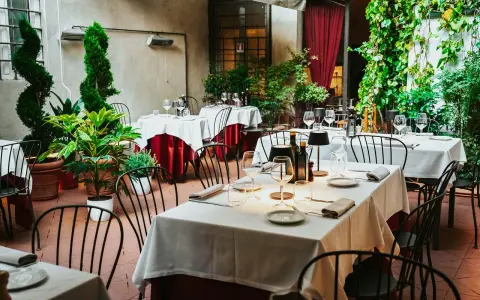 Cortile di un ristorante, tavoli apparecchiati con tovaglie bianche e rosse, piante verdi e sedie nere. Un invito a mangia e bevi santo spirito.