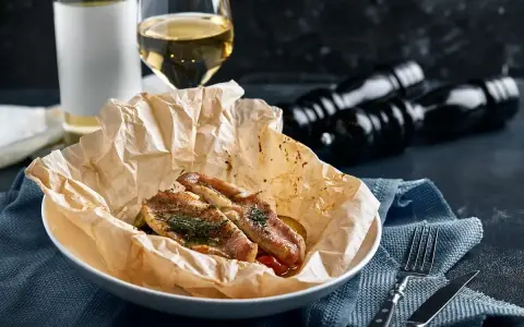 Deliziosa trota puccini cotta al forno, servita su carta da forno con erbe aromatiche. Accanto, un bicchiere di vino bianco e macinapepe.