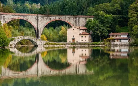 Pieve Fosciana: due ponti, uno antico e uno moderno, si specchiano in un lago calmo, con edifici storici e una foresta sullo sfondo.