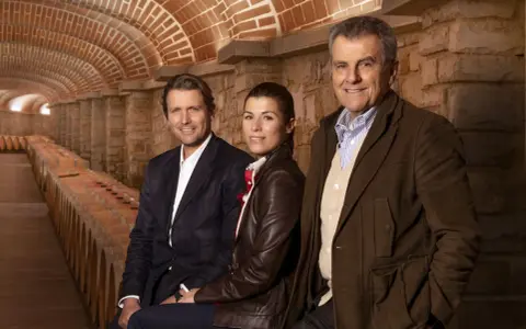 Tre persone sorridono nella cantina della Tenuta Il Borro, circondati da botti di vino e un soffitto a volta in mattoni.