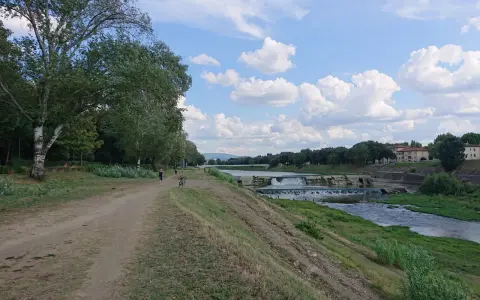 Sentiero lungo il fiume con alberi e il masso della Gonfolina in lontananza. Cielo azzurro con nuvole bianche.