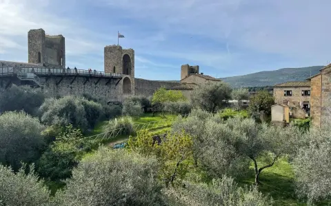 Le mura di Monteriggioni, Siena, dominano un paesaggio toscano con ulivi e colline. Un ponte pedonale attraversa la fortificazione.