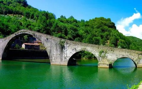 Il Ponte del Diavolo, con i suoi archi in pietra, attraversa un fiume verde smeraldo, circondato da colline lussureggianti e un cielo azzurro.