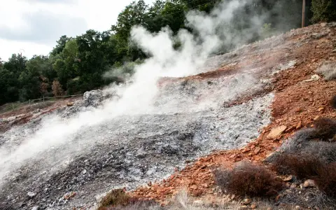 Fumarole attive nel Parco delle Biancane, con vapore che sale da un terreno roccioso e argilloso, circondato da vegetazione e alberi.