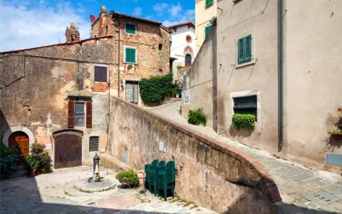 Un vicolo pittoresco a Castagneto Carducci, con case antiche, un fontanello e sedie verdi impilate.