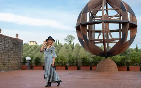 Donna con cappello nero ammira l'Uomo Vitruviano in una sfera di legno, un'opera iconica da vedere a Vinci, con colline verdi sullo sfondo.