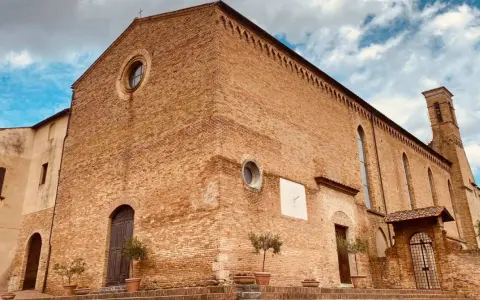 La **chiesa di Sant'Agostino** a San Gimignano, con la sua facciata in mattoni rossi e il campanile svettante, si staglia contro un cielo azzurro e nuvoloso.