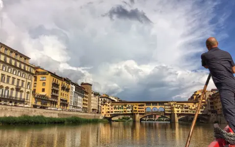 Un uomo su una barca a remi naviga lungo l'Arno, ammirando Ponte Vecchio e gli edifici storici di Firenze.