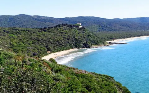 Vista panoramica delle bandite di Scarlino, con una spiaggia dorata bagnata da un mare azzurro e una villa in cima a una collina verde.