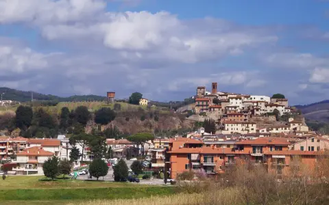 Paesaggio collinare con borgo medievale in cima, case moderne ai piedi. Si intravede un serbatoio d'acqua, vigneti e il **limite** del paese.