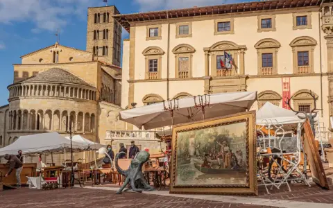 Mercatino dell'antiquariato in piazza grande, con opere d'arte, sculture e mobili antichi esposti sotto il sole.