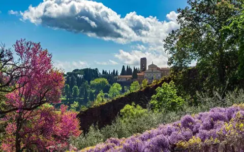 Giardini fioriti con glicini viola e alberi rosa, sullo sfondo un edificio storico con torre: curiosità da vedere a Firenze.