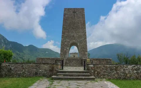 Monumento a Sant'Anna di Stazzema, cosa vedere: un imponente torrione in pietra con arco, che custodisce una tomba, circondato da mura e montagne.