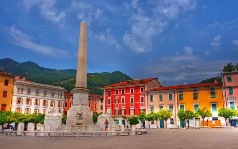 Piazza con obelisco e leoni, edifici colorati e montagne sullo sfondo. Un'idea di cosa vedere a Marina di Massa.