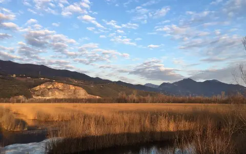 Acque calme del lago di Porta riflettono un cielo azzurro con nuvole bianche, circondato da canneti dorati e montagne all'orizzonte.