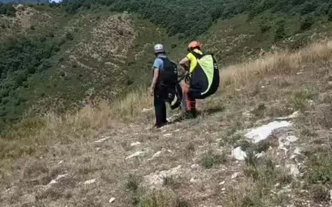 Due parapendisti si preparano al decollo sulla **Valle del Serchio**, con un paesaggio verdeggiante alle spalle.