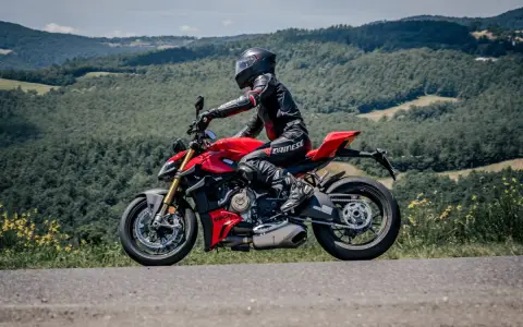 Motociclista in tuta rossa e nera sfreccia su una Ducati rossa lungo il passo del giogo, tra colline verdi e cielo azzurro.