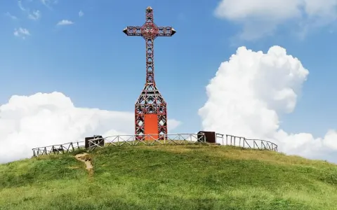 Croce metallica rossa sulla cima di una collina erbosa, con un sentiero che sale. Il **Pratomagno Arezzo** domina il paesaggio sotto un cielo azzurro con nuvole bianche.