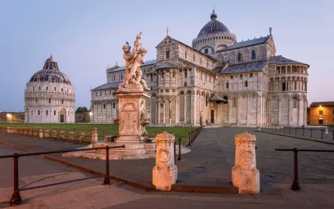 Il **Battistero di Pisa** e il Duomo illuminati dalla luce dell'alba. Una fontana con putti al centro della piazza.