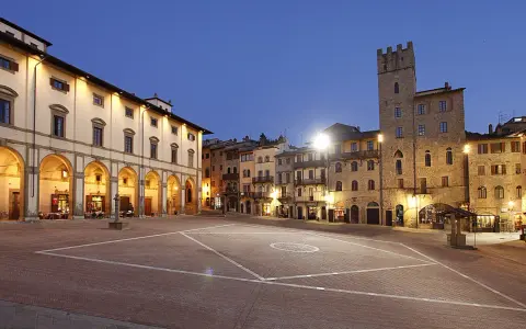 Piazza illuminata di Arezzo al crepuscolo, con portici, negozi e una torre medievale. Qui la vita è bella.