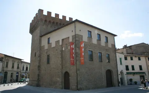 Il cassero di Montevarchi, un'antica torre con merlature, si erge imponente accanto a edifici storici, con bandiere rosse che adornano la facciata.