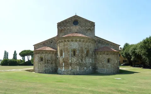 La Basilica di San Piero a Grado, con le sue absidi romaniche e il prato verde circostante, si staglia contro un cielo limpido.