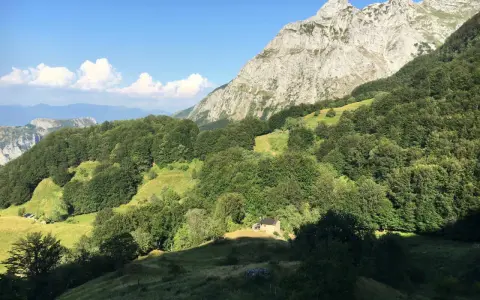 Un piccolo rifugio solitario ai piedi del **Puntato Apuane**, circondato da boschi verdi e montagne imponenti sotto un cielo azzurro.